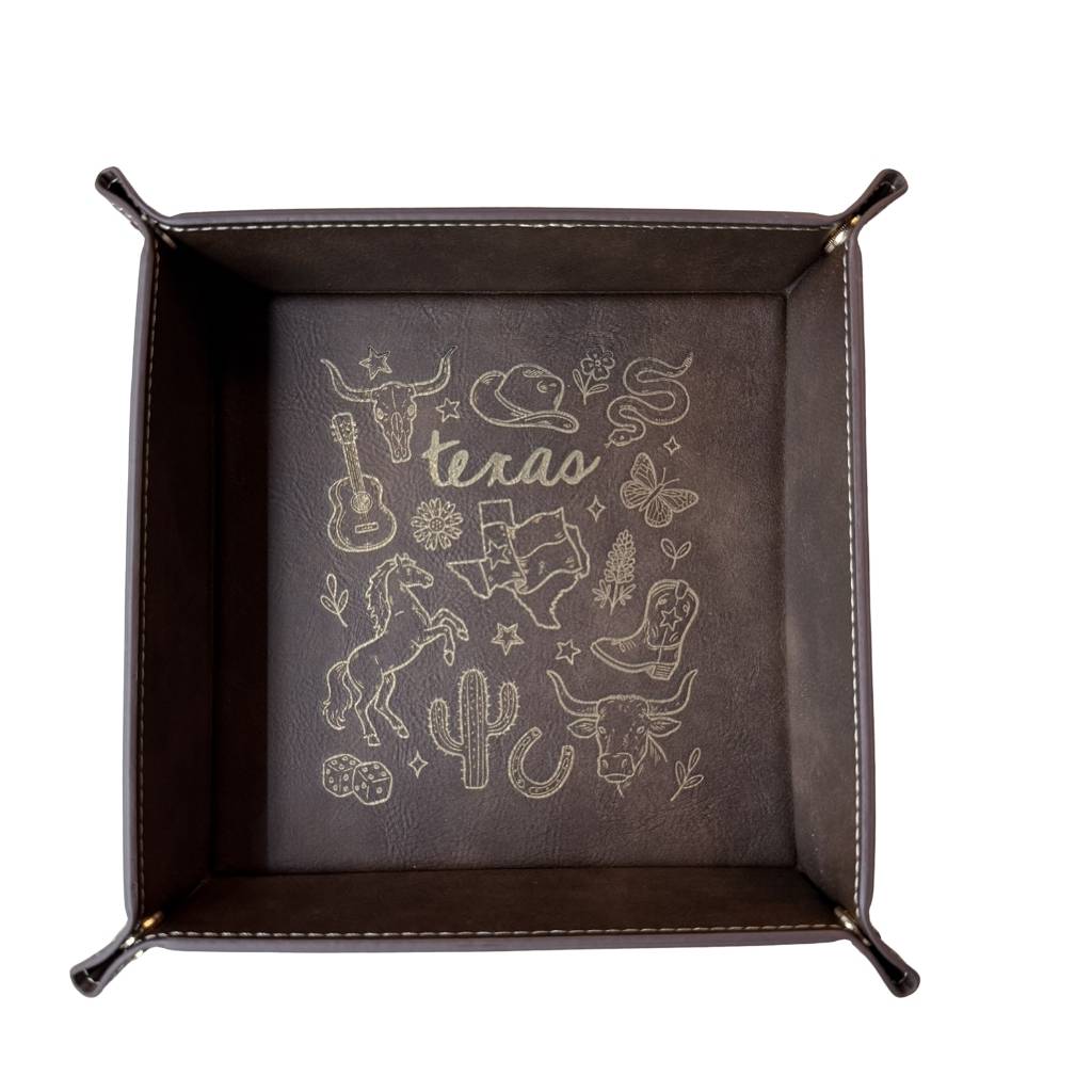 Dark brown leatherette valet tray with laser-engraved Texas illustration including longhorn skull, cowboy hat, cowboy boots, cactus, and guitar, folded open on a light wood surface