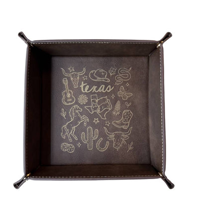 Dark brown leatherette valet tray with laser-engraved Texas illustration including longhorn skull, cowboy hat, cowboy boots, cactus, and guitar, folded open on a light wood surface