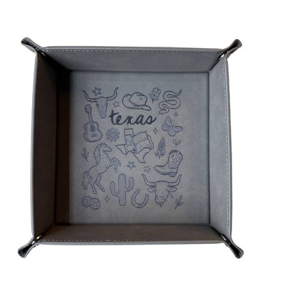 Gray leatherette valet tray with laser-engraved Texas illustration including longhorn skull, cowboy hat, cowboy boots, cactus, and guitar, folded open on a light wood surface