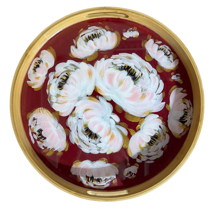 A round decorative tray with a gold-toned rim, featuring a painted floral design of large white and pale pink blossoms on a deep red background.