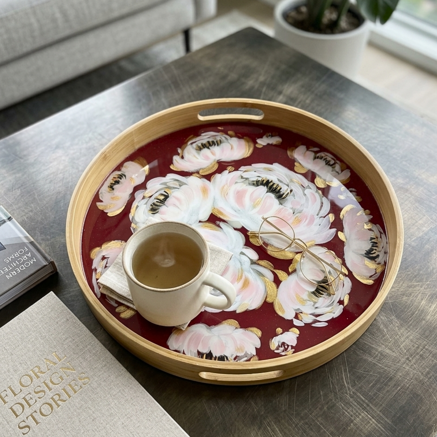 A round wooden serving tray with a deep red floral print and cutout handles, styled on a dark wood table with a white mug, eyeglasses, and a book nearby.