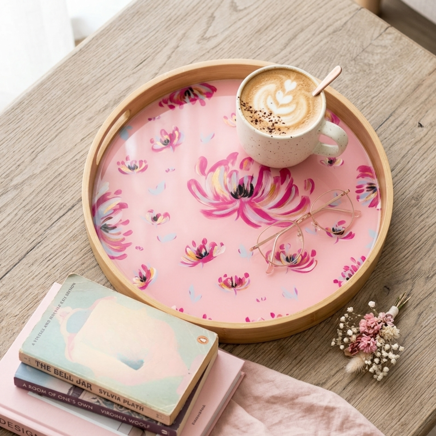 A round wooden tray with a pink floral printed liner sits on a light wood table, styled with a cup of latte, eyeglasses, books, and a small dried flower