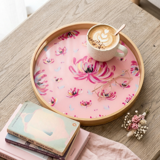 A round wooden tray with a pink floral printed liner sits on a light wood table, styled with a cup of latte, eyeglasses, books, and a small dried flower