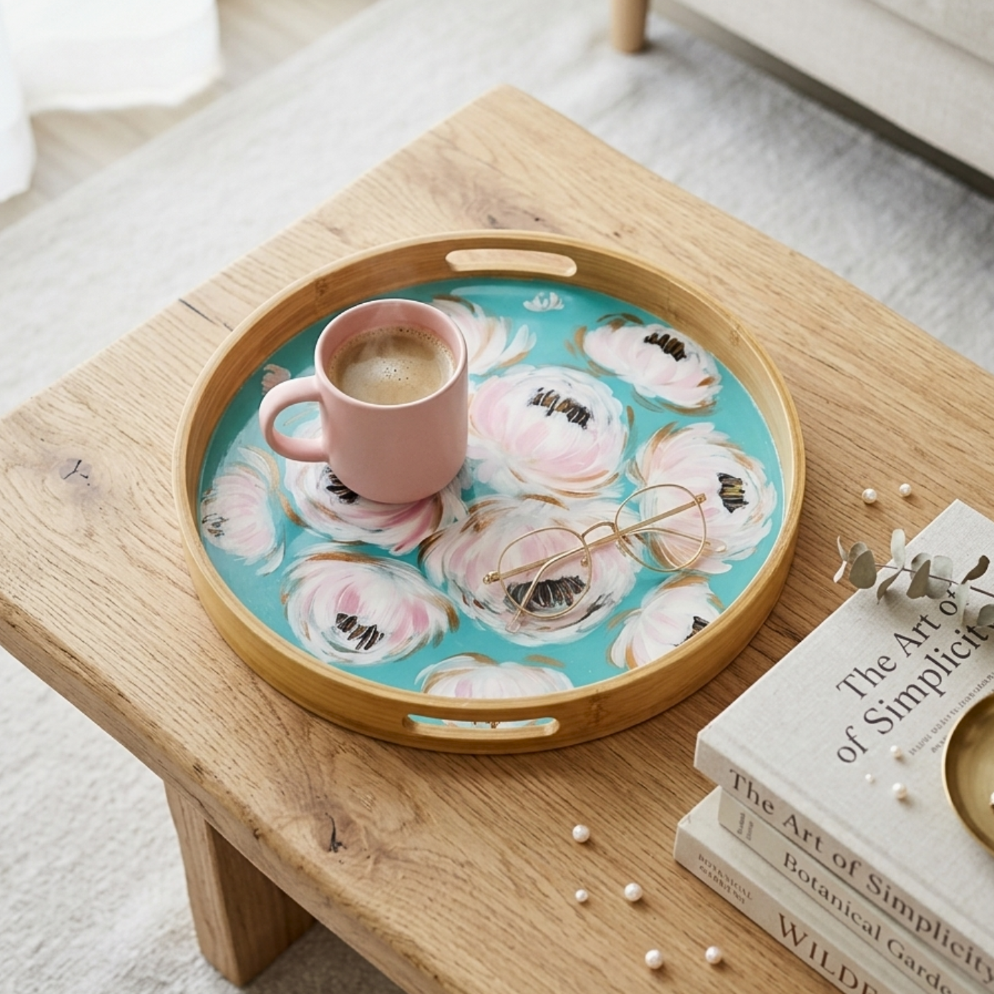 A round bamboo serving tray with a turquoise floral print sits on a light wood table, styled with a pink mug, a pair of eyeglasses, and books in a bright indoor setting.