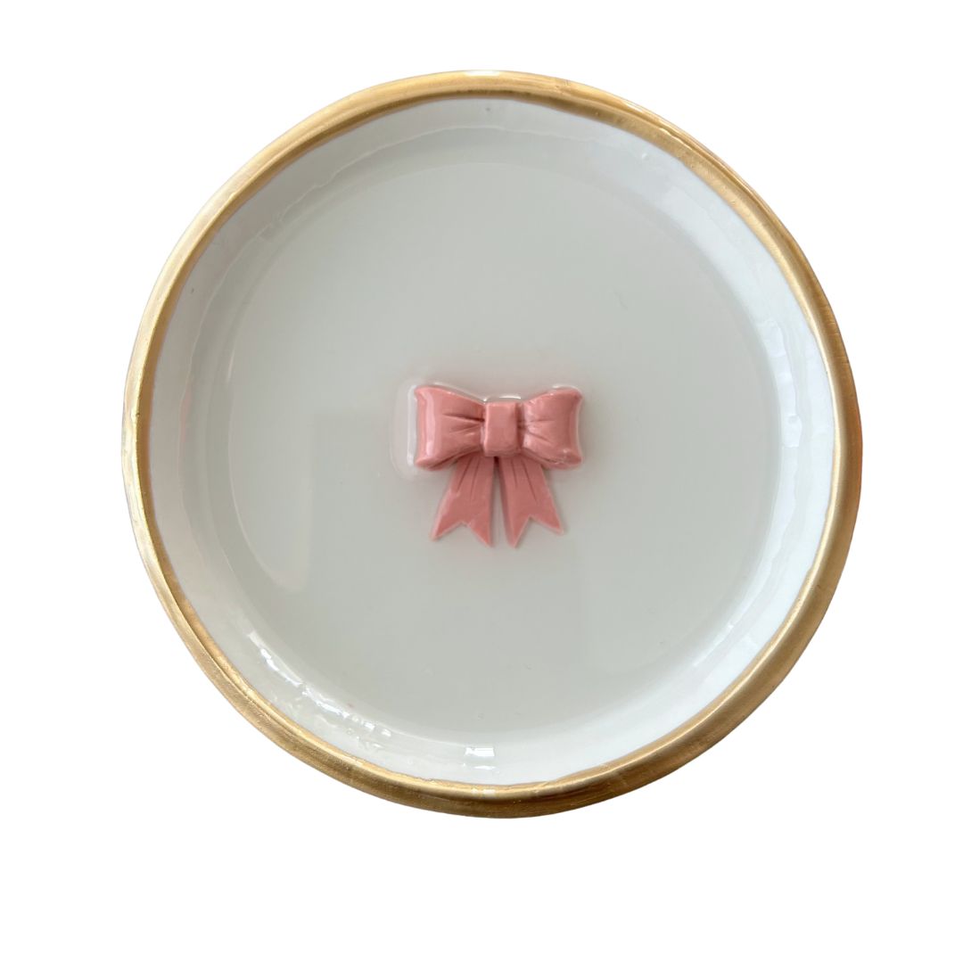 A handcrafted clay ring dish with a pink bow detail, placed on a white surface with a gold rim.