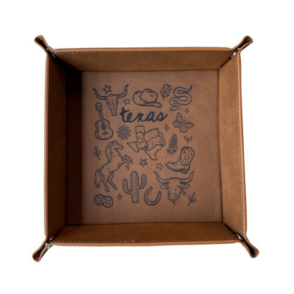 Tan caramel leatherette valet tray with laser-engraved Texas illustration including longhorn skull, cowboy hat, cowboy boots, cactus, and guitar, folded open on a light wood surface