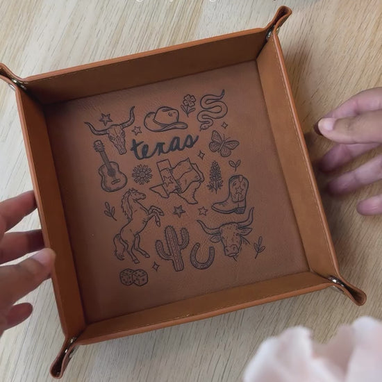 leatherette valet tray with laser-engraved Texas illustration including longhorn skull, cowboy hat, cowboy boots, cactus, and guitar, folded open on a light wood surface